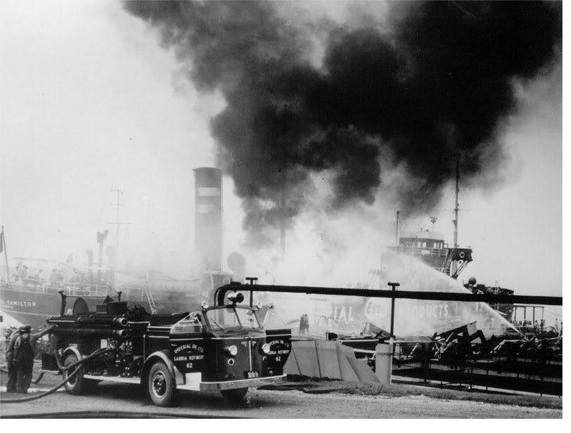 Thick black smoke pouring from a burning tanker at a dock, with firefighters and hoses in the foreground.