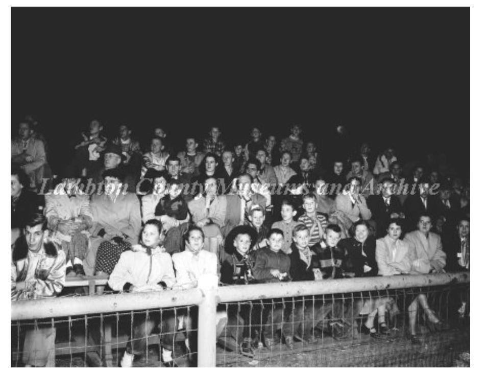 A night photograph of a crowd packed into bleachers behind a wire fence, coats and headscarves, all faces turned to the field.