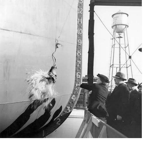 A person in a fur stole swings a bottle against the bow of a ship beside the painted draft numbers.