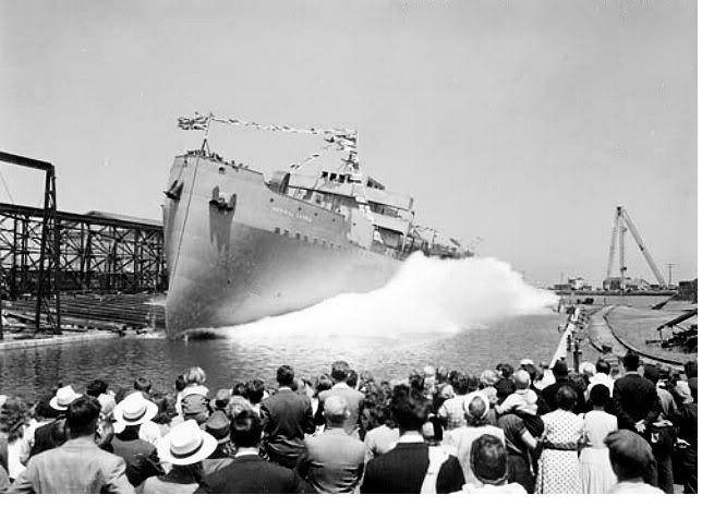 A ship's hull sliding into the water, throwing up spray, watched by a large crowd in hats.