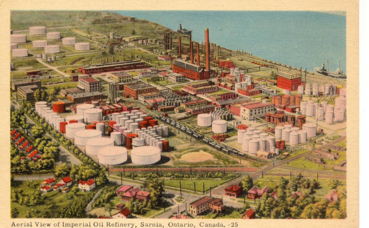 A tinted aerial postcard of an oil refinery: rows of white storage tanks, red brick buildings, tall stacks, rail sidings, a ship at the dock.