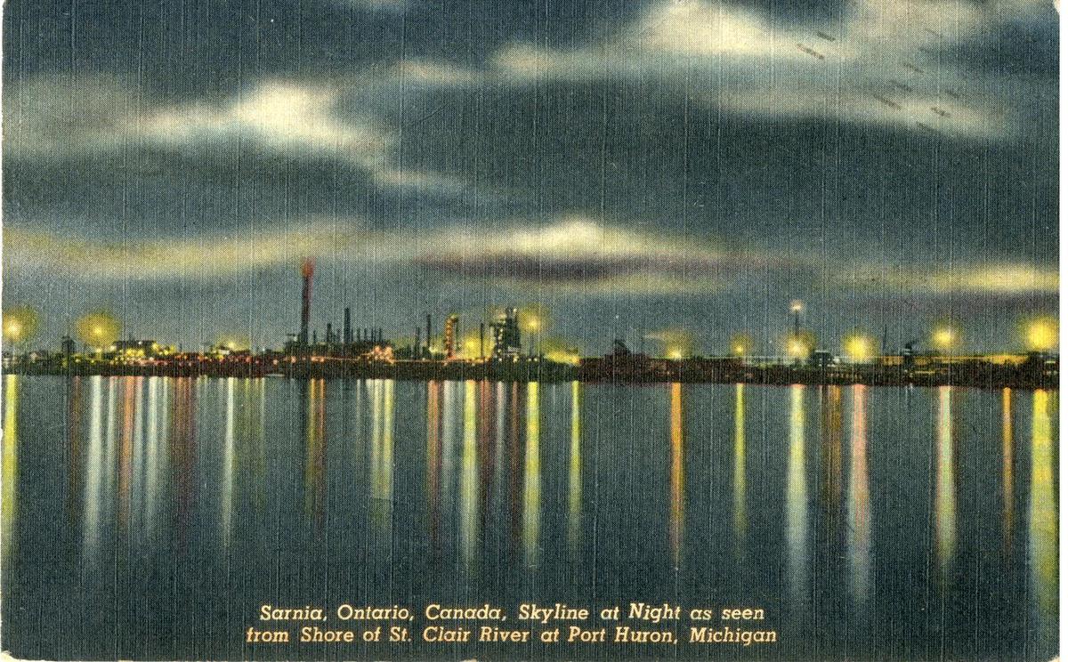 A night postcard: refinery lights along a dark shore, reflected in long streaks across the water.