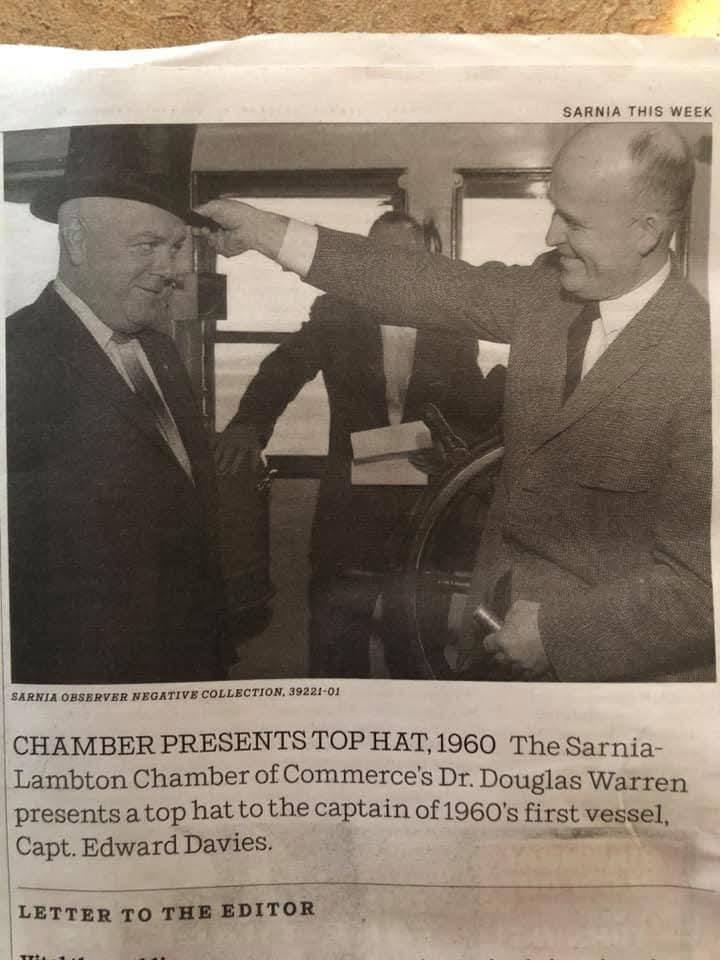Newspaper photograph: one man places a top hat on another man's head beside a ship's wheel.