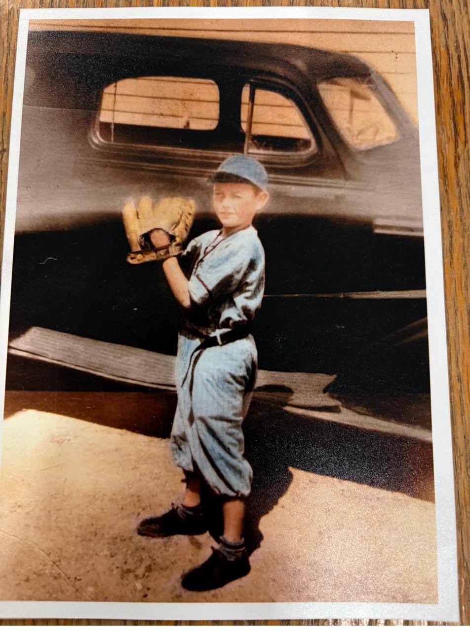 A boy in a pale baseball uniform and cap holds up a catcher's glove in front of an old sedan.