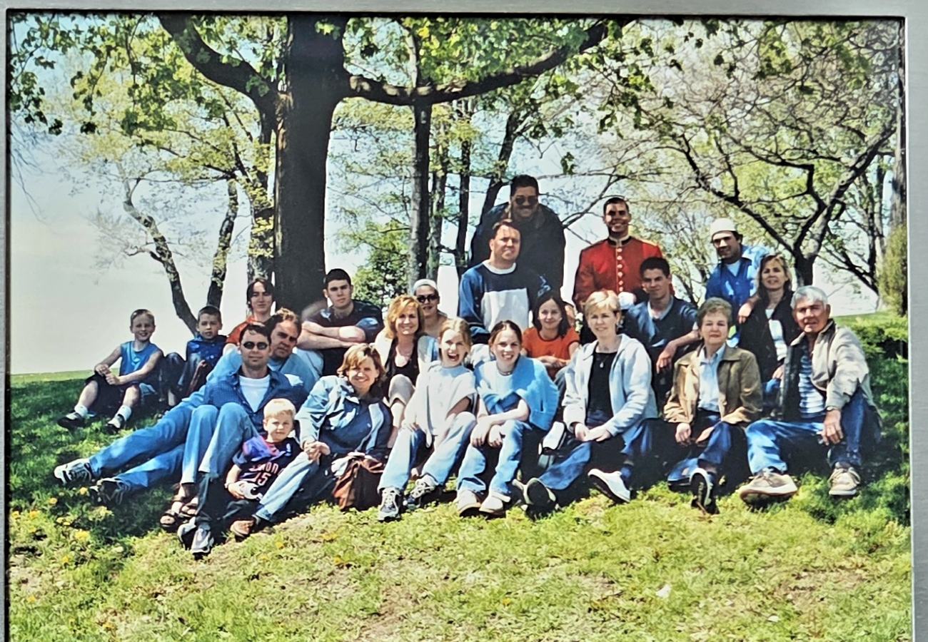 An extended family of about thirty people of all ages seated together on a grassy slope beneath tall trees.
