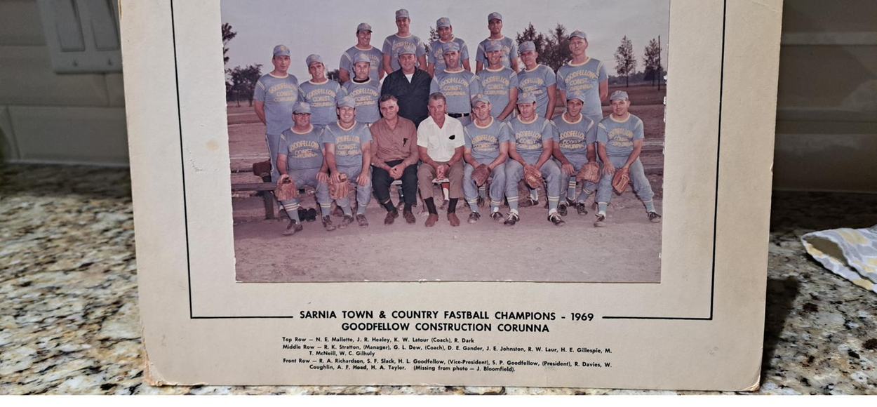 A mounted team photograph of a fastball squad in pale uniforms, with a printed caption and name list below.