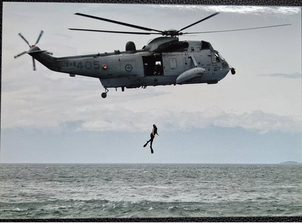 A Sea King helicopter over open water with a diver falling from the door toward the sea.