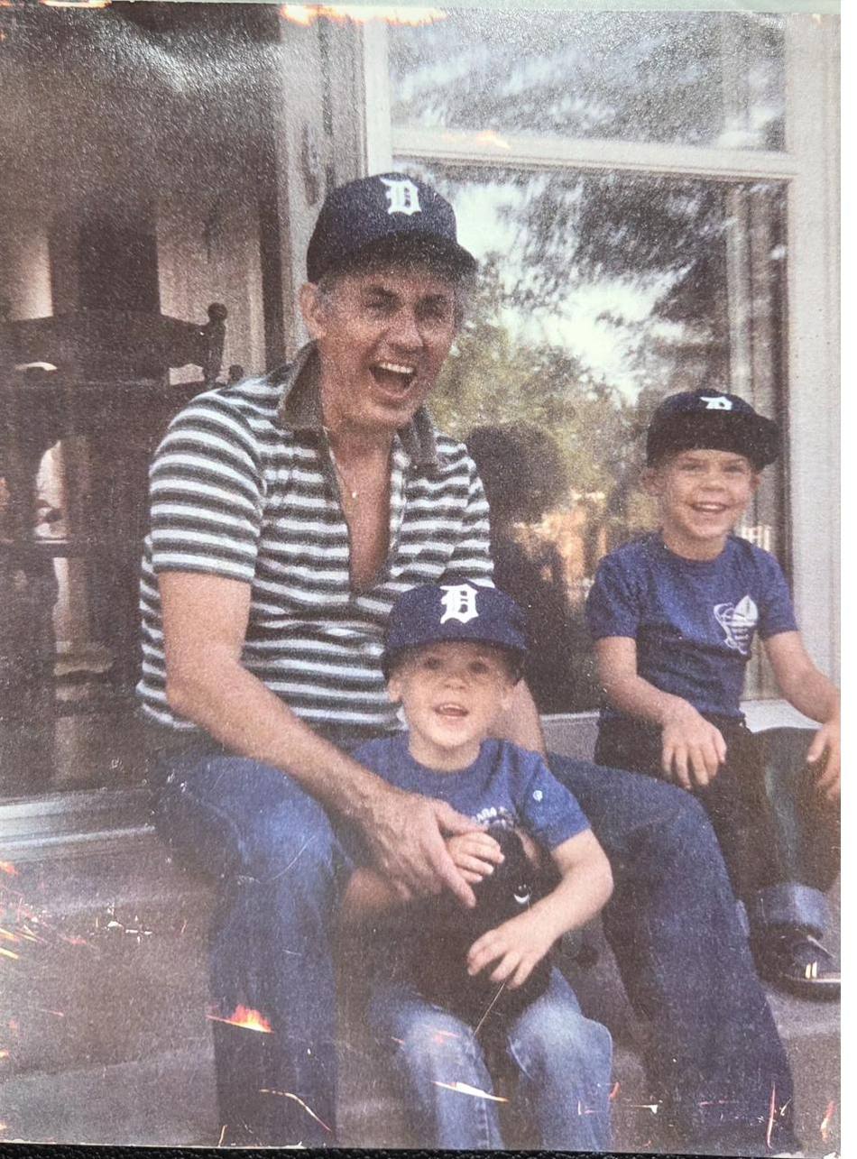 A laughing man in a Detroit Tigers cap on a porch step with two small boys in matching caps.